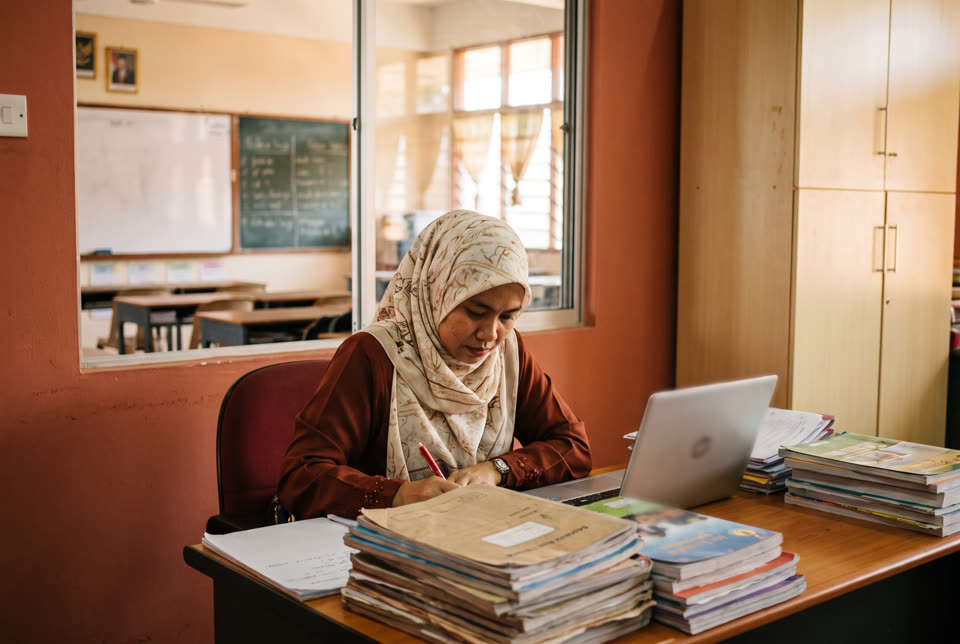 Seorang guru wanita bertudung menyemak kerja di bilik guru dengan komputer riba.