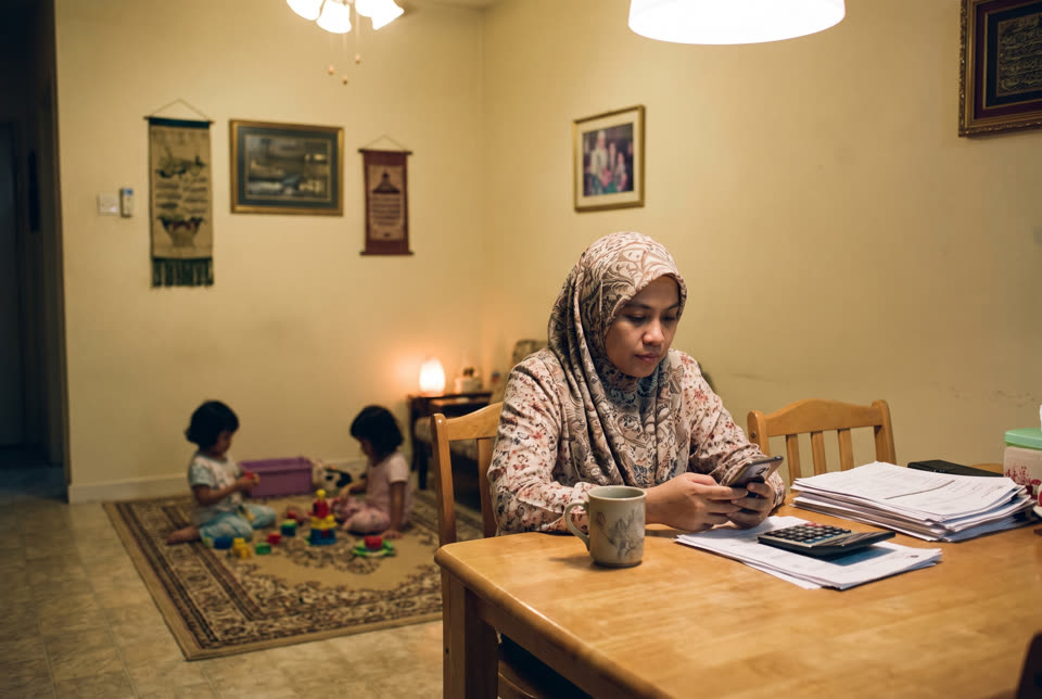 Seorang ibu di meja makan menyemak bil dan telefon, anak-anak bermain di belakang.